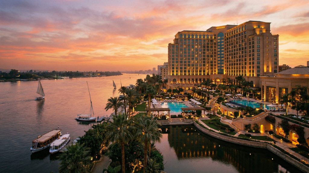 Vue du Four Seasons Cairo at Nile Plaza au coucher du soleil, surplombant le Nil avec jardins luxuriants, piscine et bateaux traditionnels.