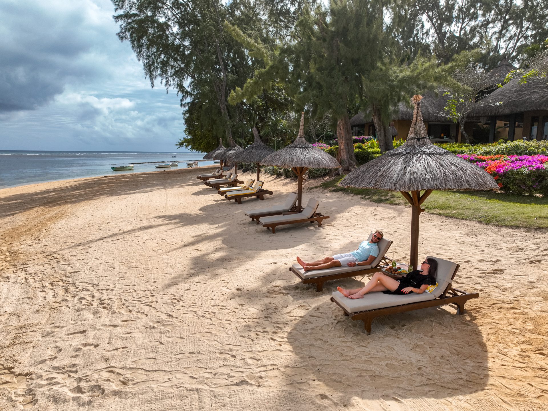 Plage du The Oberoi Beach Resort Mauritius, couple se relaxant sur des transats sous des parasols en chaume face au lagon turquoise