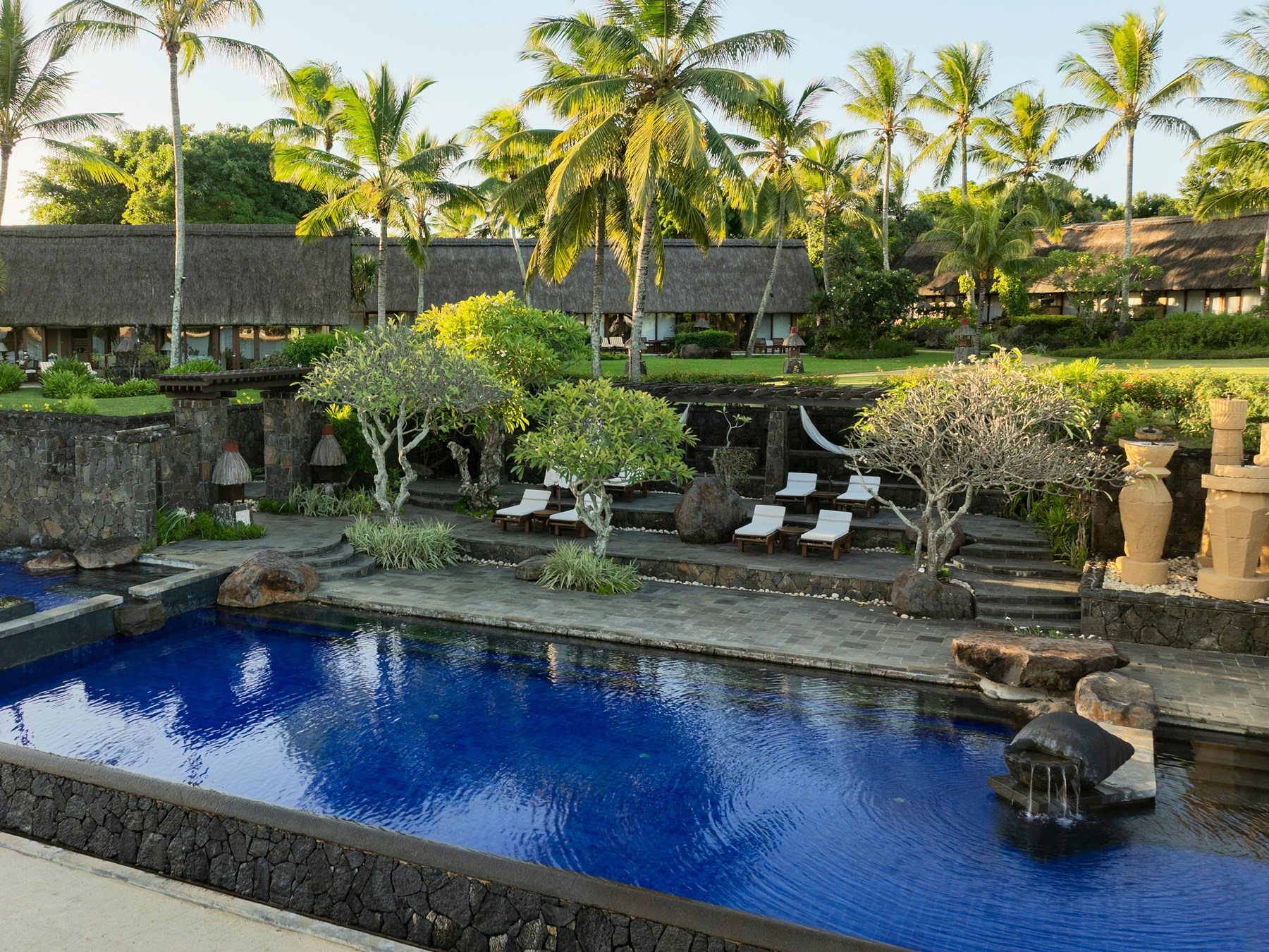 Piscine du The Oberoi Beach Resort Mauritius, bassin bleu entouré de jardins tropicaux, palmiers et architecture mauricienne en pierre naturelle.