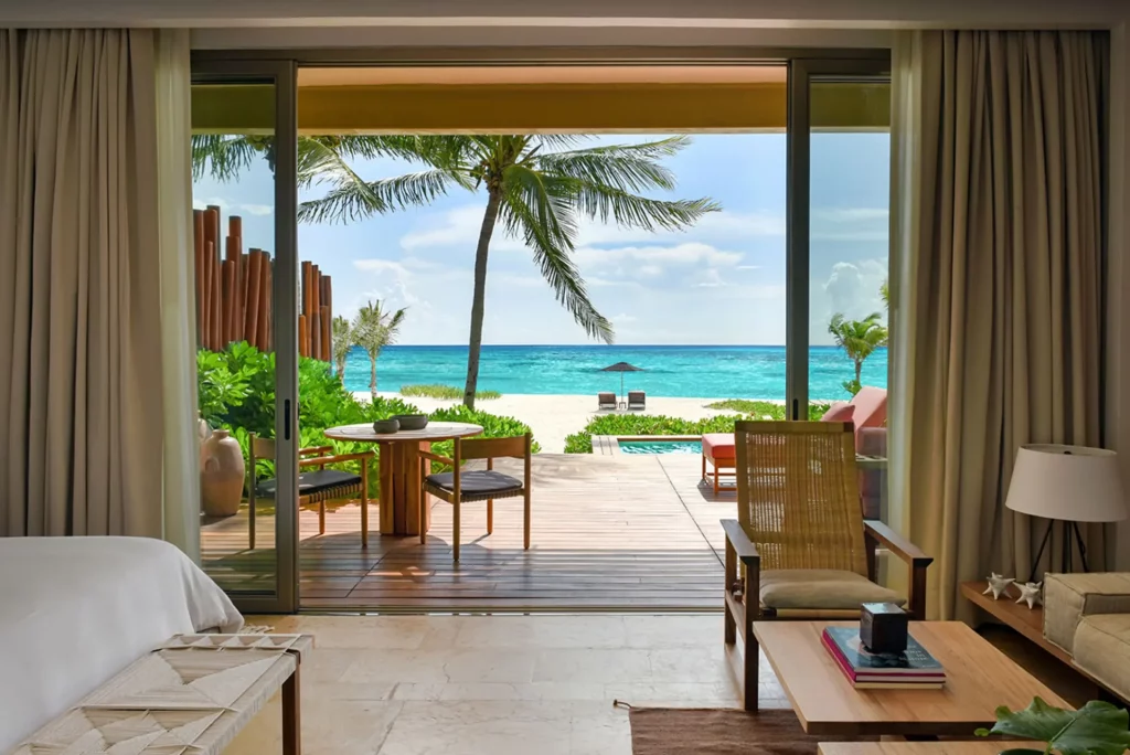 Vue depuis l'intérieur de la Beachfront Studio Suite du Rosewood Mayakoba, baies vitrées ouvertes sur la terrasse, palmier et mer des Caraïbes turquoise, Riviera Maya Mexique